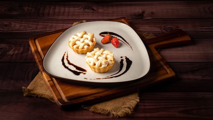 Two small tarts with cream filling are on a triangular plate, garnished with chocolate sauce drizzles and two strawberries.