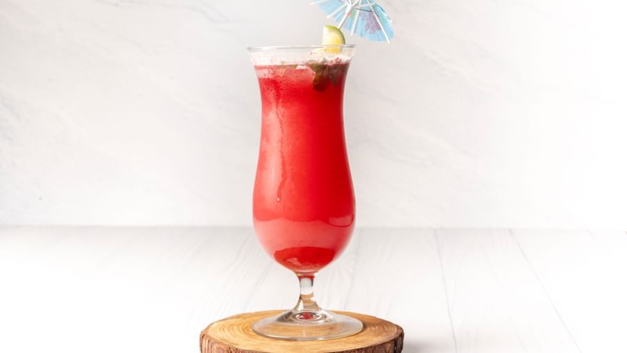 A tall glass of beverage with a straw and a small blue umbrella sits on a wooden coaster.