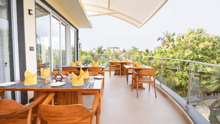 An open-air dining area with wooden tables and chairs, set with yellow napkins and glassware, overlooking lush greenery with a modern glass railing and a curved white canopy - MGM Beach Resorts