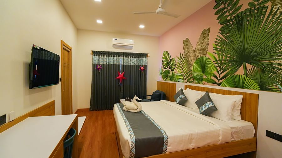 Image of bedroom under warm lighting featuring a plush bed with a TV in front of it and a window on the other side