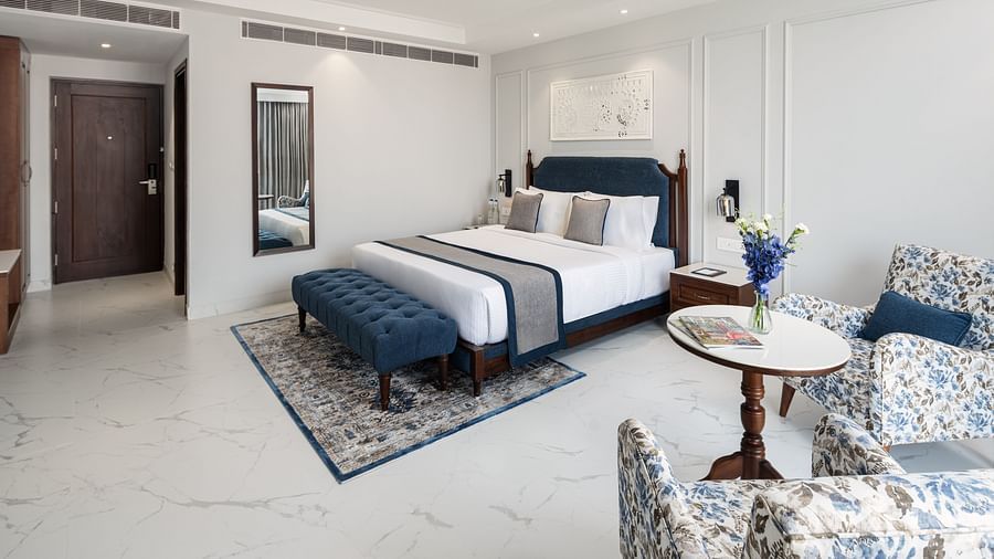 A hotel room with a large bed, upholstered chairs, and a round table with a flower vase - MJ Sarovar Portico, Haridwar
