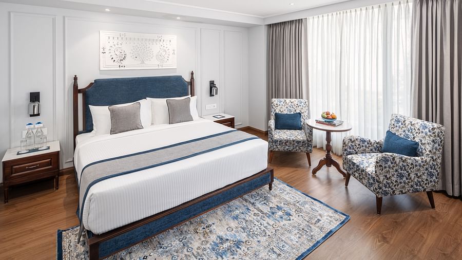 A room with a large double bed, wooden headboard, nightstand, and upholstered sofas - MJ Sarovar Portico, Haridwar