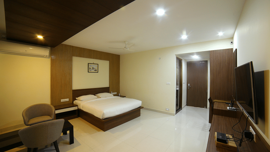 A room featuring a well made bed sofa chairs a TV and a wall painting - Mysore Gate Hotel Bangalore