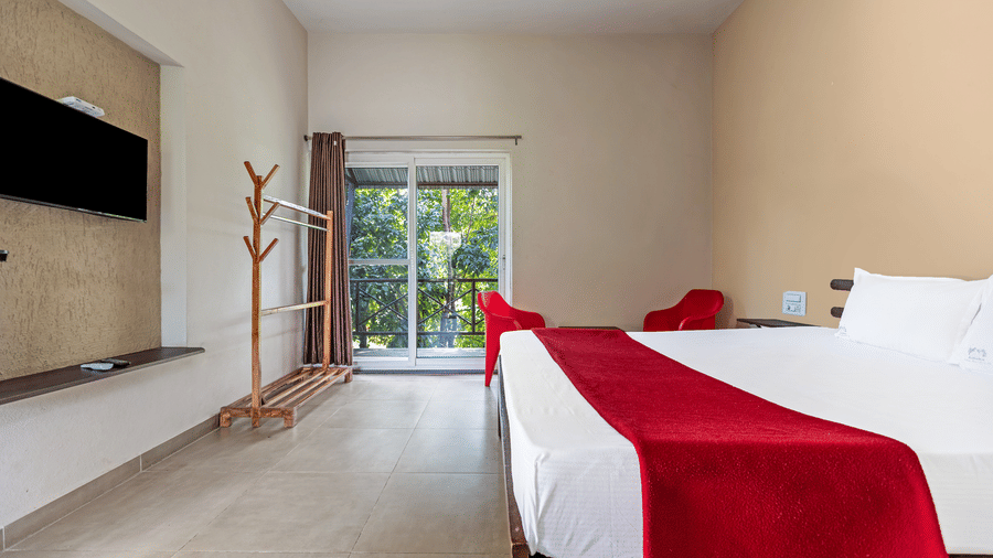 Interior view of a hotel room showing a wall-mounted TV, wooden clothes rack, and glass doors leading to a balcony - Nature Trails Ashoka Resort Hampi
