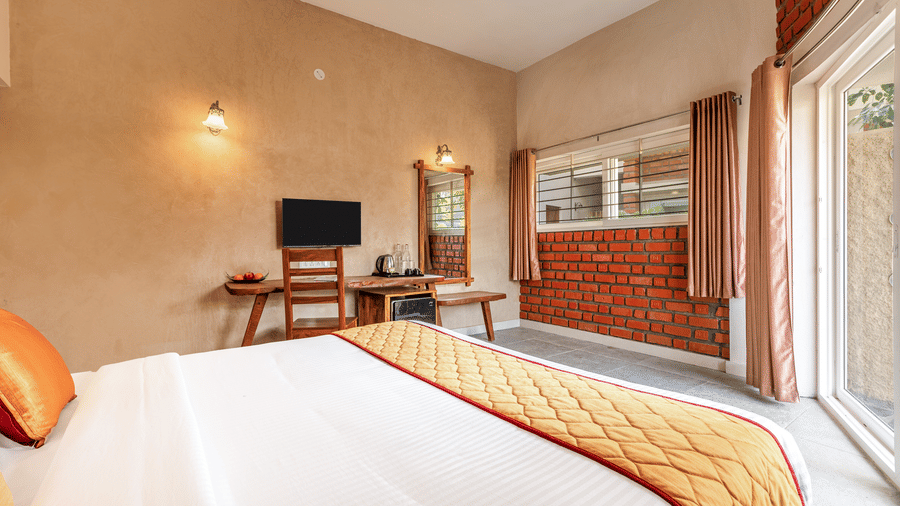 Bedroom interior showing the orange bed runner and floor-to-ceiling windows letting in natural light - Nature Trails Ashoka Resort Hampi