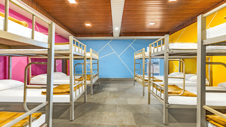 Nature Trails Kudalika 20 dom room full view with rows of metal bunk beds, white bedding, mustard blankets, pink yellow and blue geometric walls and wood-panelled ceiling.