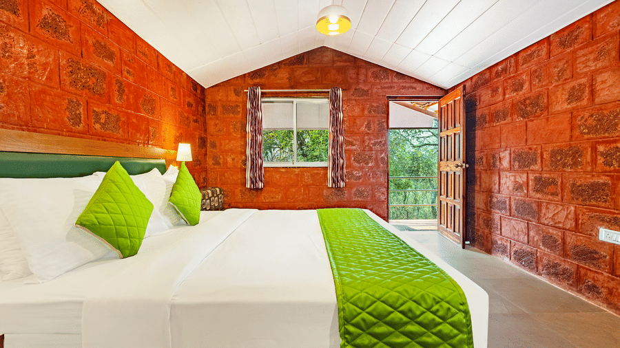 Nature Trails Kudalika premium heritage room with double bed, green bedding, red laterite walls, window with purple curtains and open wooden door to balcony.