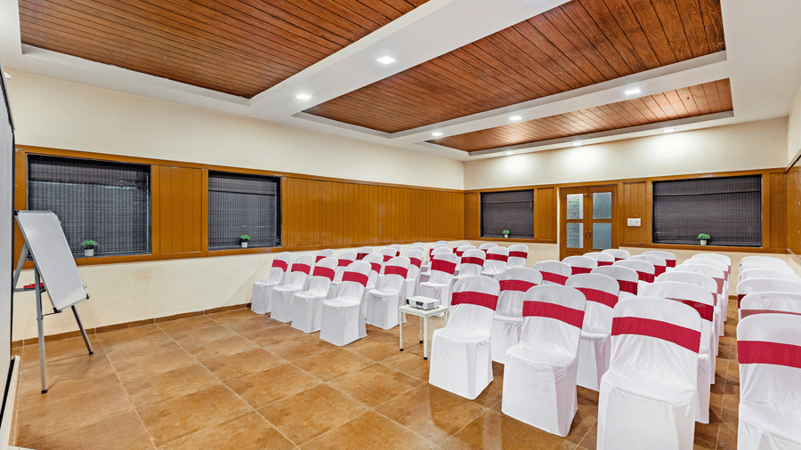 Nature Trails Kudalika conference hall with theatre-style white and red chairs, projector on central table, flipchart stand and wooden panelled walls.