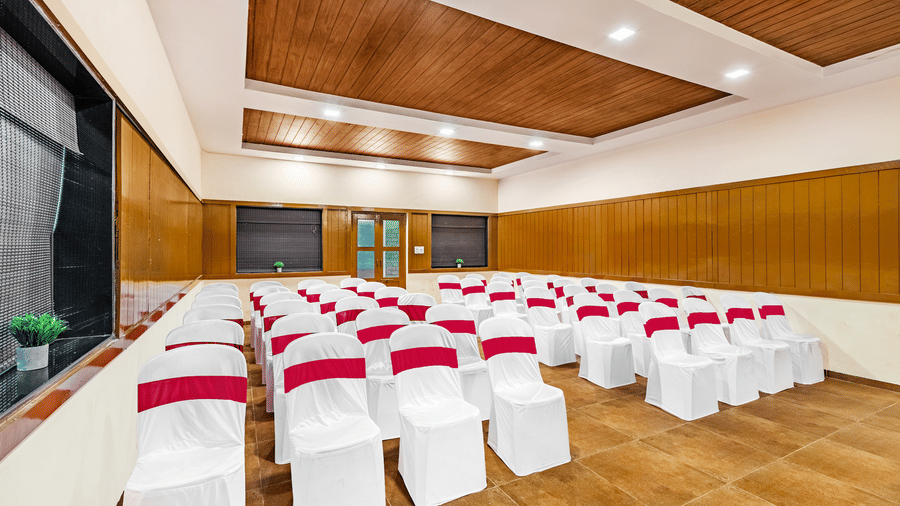 Nature Trails Kudalika conference hall side view showing theatre-style white and red chairs, wooden wall panelling, roller blinds and coffered wood ceiling.