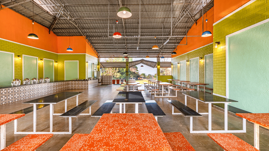 Nature Trails Kudalika dining hall showing bench tables with orange covers, tiled buffet counter with steel vessels, coloured pendant lights and open garden view.