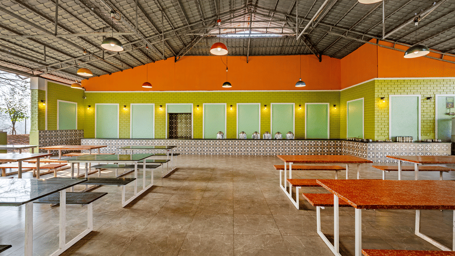 Nature Trails Kudalika dining hall with mixed bench tables, tiled buffet counter with steel vessels, green brick walls, orange upper walls and coloured pendant lights.