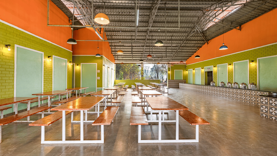 Nature Trails Kudalika dining hall long view with red bench tables, tiled buffet counter with steel vessels on the right, green brick walls and coloured pendant lights.