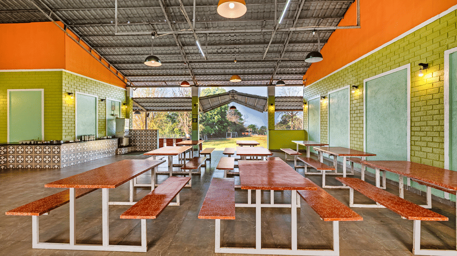 Nature Trails Kudalika dining hall with red-topped bench tables, tiled buffet counter on the left, green brick walls, coloured pendant lights and open garden view.