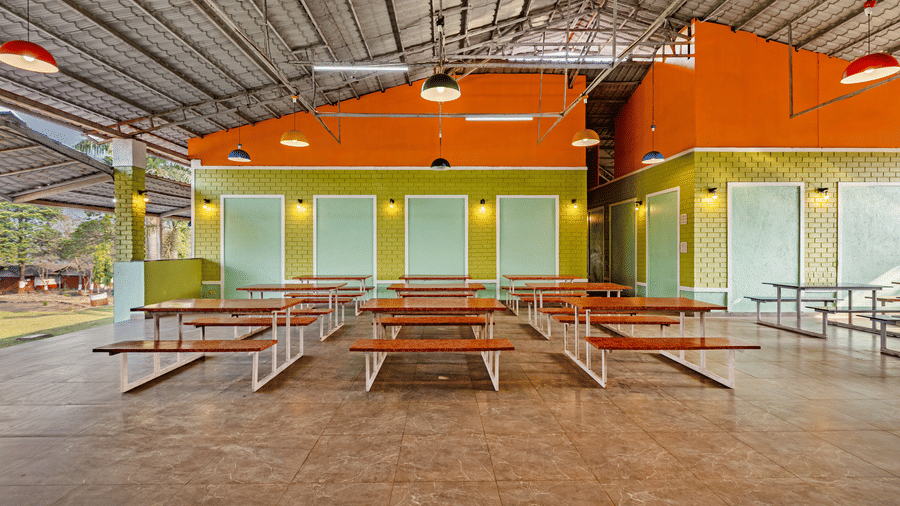 Nature Trails Kudalika open dining hall with red-topped bench tables, green brick walls, orange upper walls, coloured pendant lights and open-air sides.