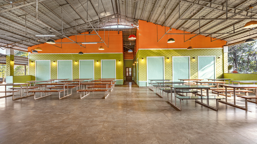 Nature Trails Kudalika open dining hall with bench seating, green brick walls, orange upper walls, colourful pendant lights and corrugated metal roof.