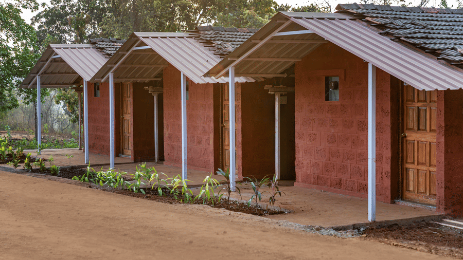 Nature Trails Kudalika laterite stone cottages with white corrugated porch roofs, wooden doors, white pillars and young plants along a red dirt pathway.