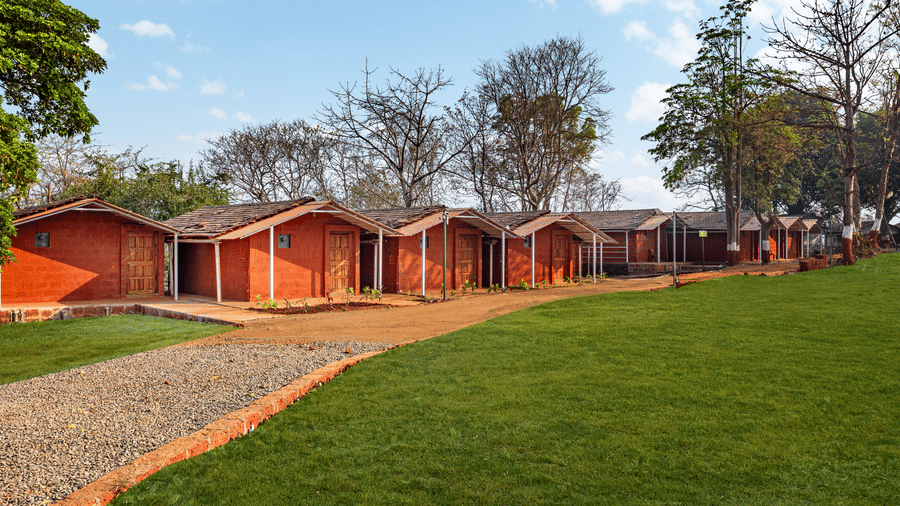 Nature Trails Kudalika row of red laterite cottages with wooden doors, tiled roofs and small verandahs, set alongside a green lawn with a gravel pathway border.