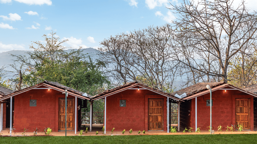 Nature Trails Kudalika three red laterite cottages with wooden panel doors, tiled roofs, wall lights and small verandahs, set against mountain and tree backdrop.