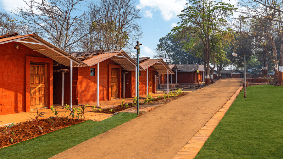 Nature Trails Kudalika row of red laterite stone cottages with wooden doors, tiled roofs, white pillars and a mud pathway flanked by green lawn and young plants.