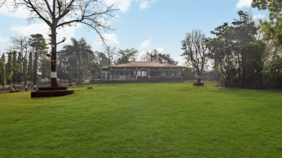 Nature Trails Kudalika expansive green lawn with a single-storey tiled roof building visible in the background, framed by bare and leafy trees under blue sky.