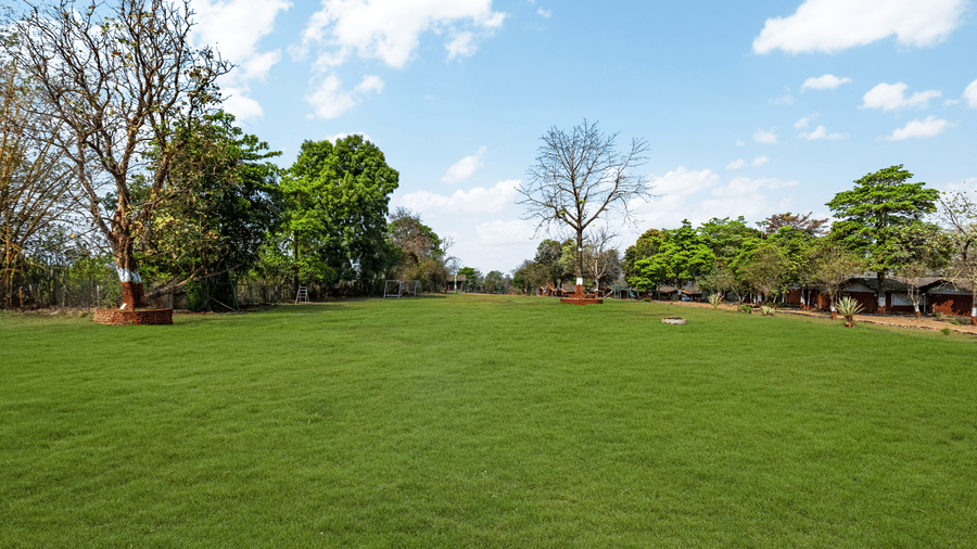 Nature Trails Kudalika wide open green lawn with surrounding trees, property structures visible in the background under a blue sky with white clouds.