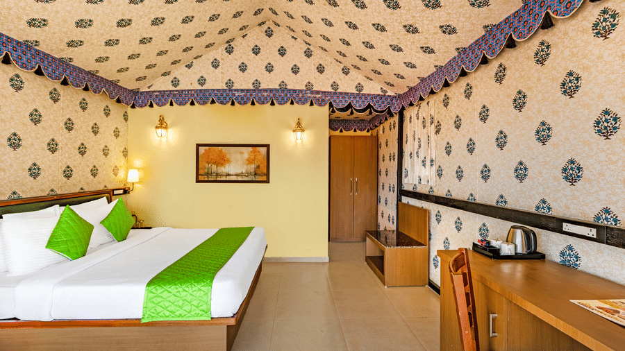 Nature Trails Kudalika premium AC tent room with double bed, green bedding, block-print fabric walls and ceiling, wooden wardrobe, work desk and brass wall lights.