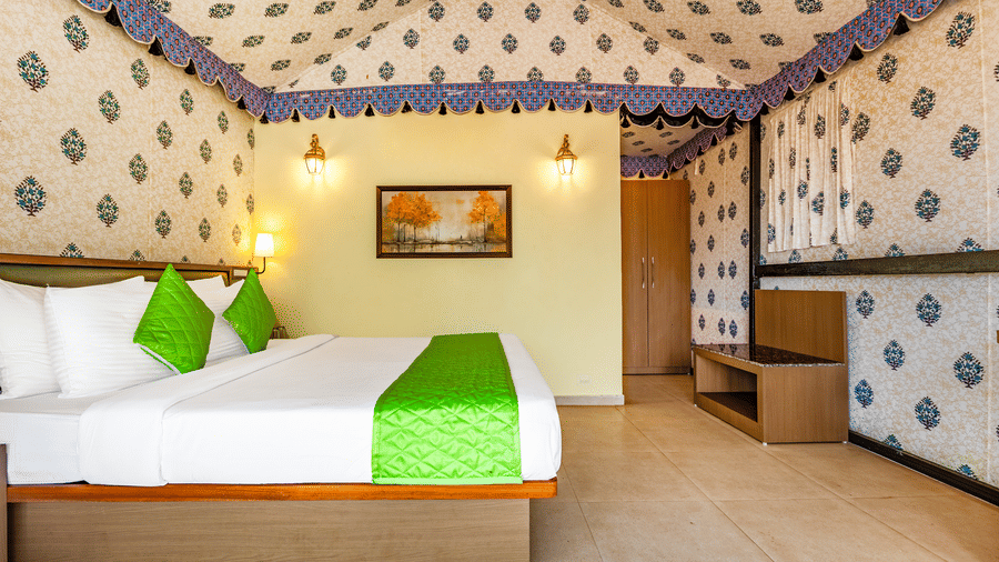 Nature Trails Kudalika premium AC tent room with double bed, green bedding, block-print fabric walls, brass wall lanterns, wooden wardrobe and framed wall painting.