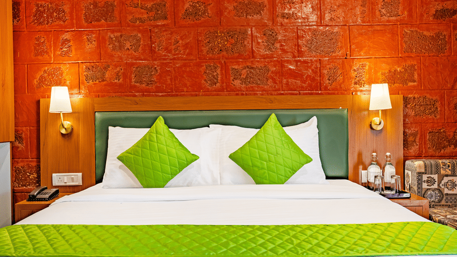 Nature Trails Kudalika premium heritage room bed front view with green quilted pillows, white bedding, green leather headboard, brass wall lamps and red laterite wall.