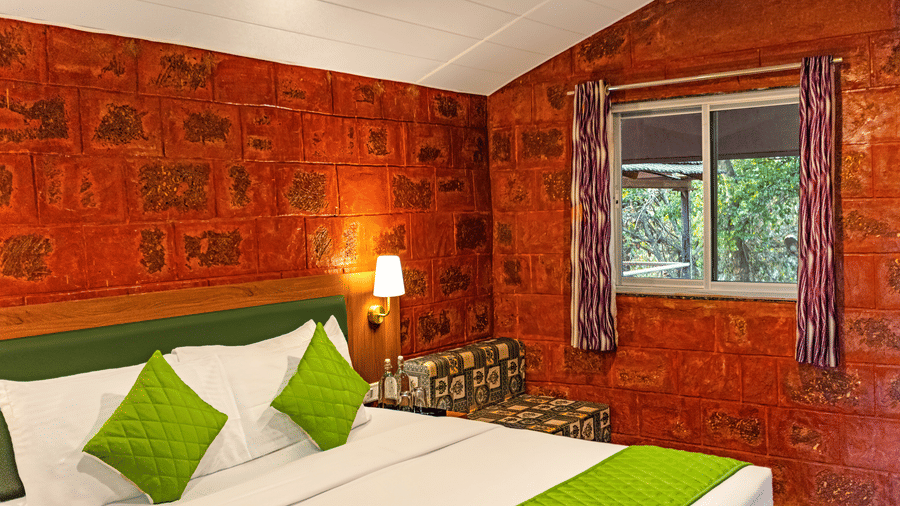 Nature Trails Kudalika premium heritage room with double bed, green bedding, exposed red laterite stone walls, window with purple curtains and garden view.
