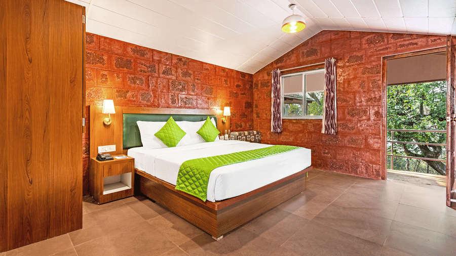 Nature Trails Kudalika premium heritage room with double bed, green bedding, red laterite stone walls, wooden wardrobe, pendant light and open balcony with tree view.