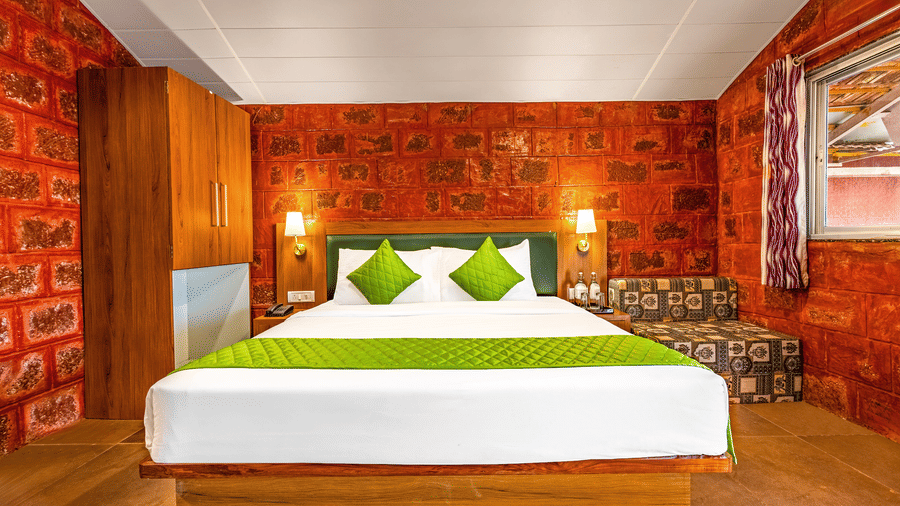 Nature Trails Kudalika premium heritage room with double bed, green bedding, red laterite stone walls, wooden wardrobe with mirror, printed bench and side window.