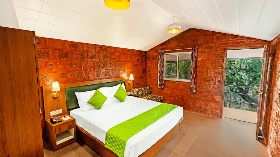 Nature Trails Kudalika premium heritage room with double bed, green bedding, red laterite stone walls, pendant lights, open balcony door with green tree view.