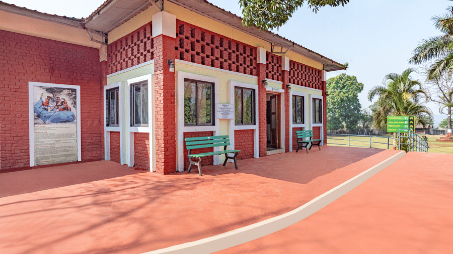 Nature Trails Kudalika reception building corner view with red laterite walls, latticed brick top, green benches, red floored pathway and directional signboard.