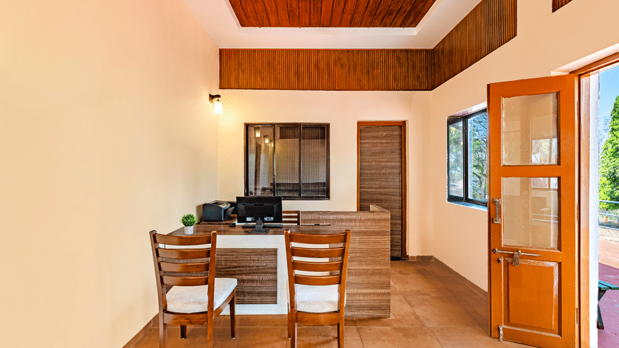 Nature Trails Kudalika reception front desk closeup with wooden counter, computer monitor, two cushioned chairs, small plant and wood-panelled coffered ceiling.
