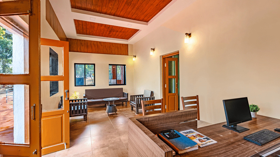 Nature Trails Kudalika reception interior with open wooden entrance door, front desk with computer, lounge seating, wood-panelled coffered ceiling and wall lights.