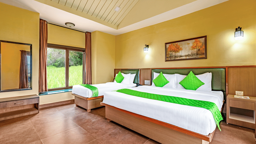 Nature Trails Kudalika river view room with two beds, green bedding, yellow walls, green headboards, wall lights, framed painting and large window with greenery view.