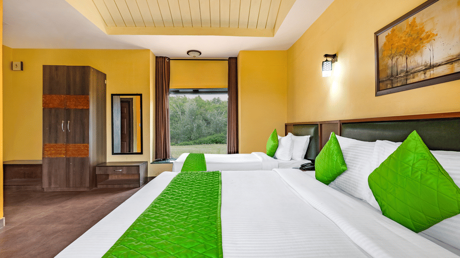Nature Trails Kudalika river view room wide view with two beds, green bedding, yellow walls, wooden wardrobe, wall light, framed painting and large nature-view window.