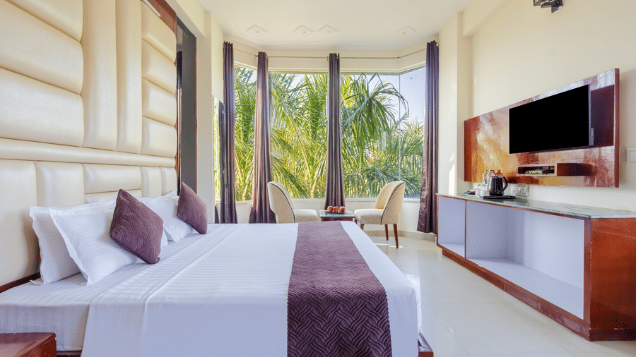 A bright hotel room featuring a large window with palm tree views and a wall-mounted television - Nature Trails Rishikesh