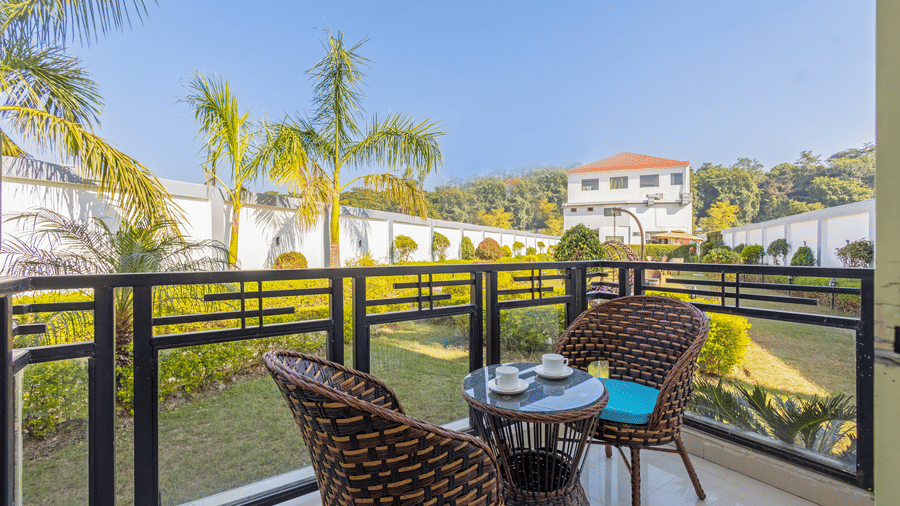 A sunny balcony with two wicker chairs and a small table overlooking a lush green garden - Nature Trails Rishikesh