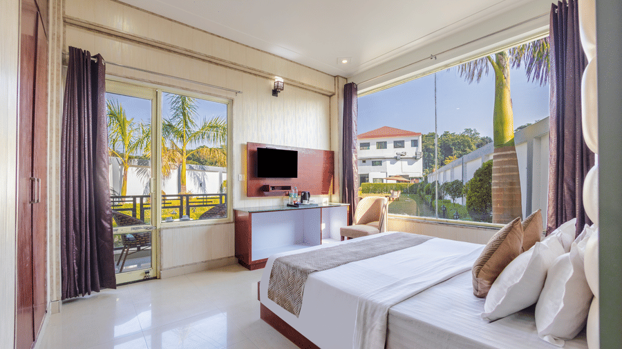 A spacious bedroom with large glass windows providing a panoramic view of the resort grounds - Nature Trails Rishikesh