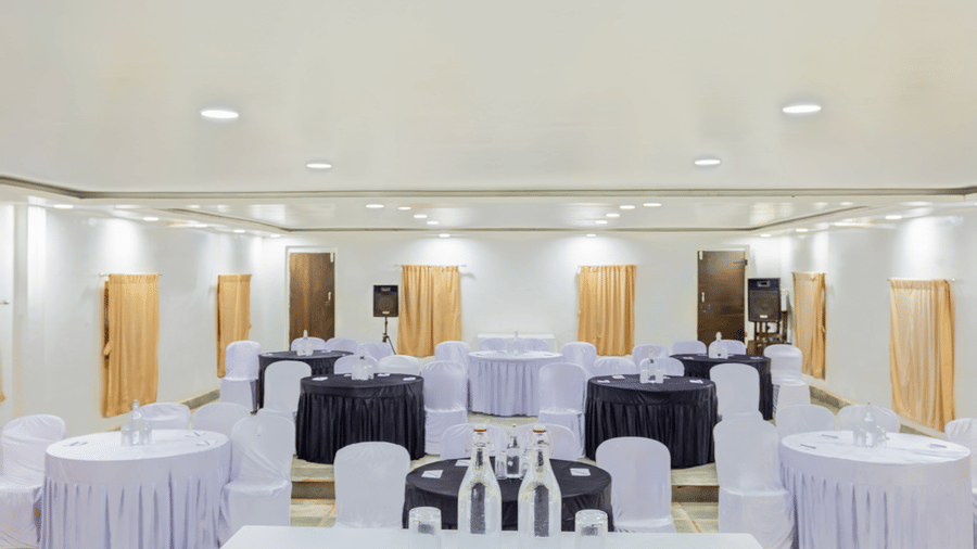 Spacious banquet hall with round tables in white and purple draping, white chair covers, warm recessed lighting and wooden partition walls in background.