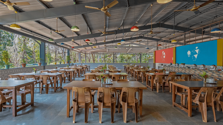 Bright open-sided resort dining area with wooden tables and chairs, transparent roof panels allowing natural light and colourful walls in background.