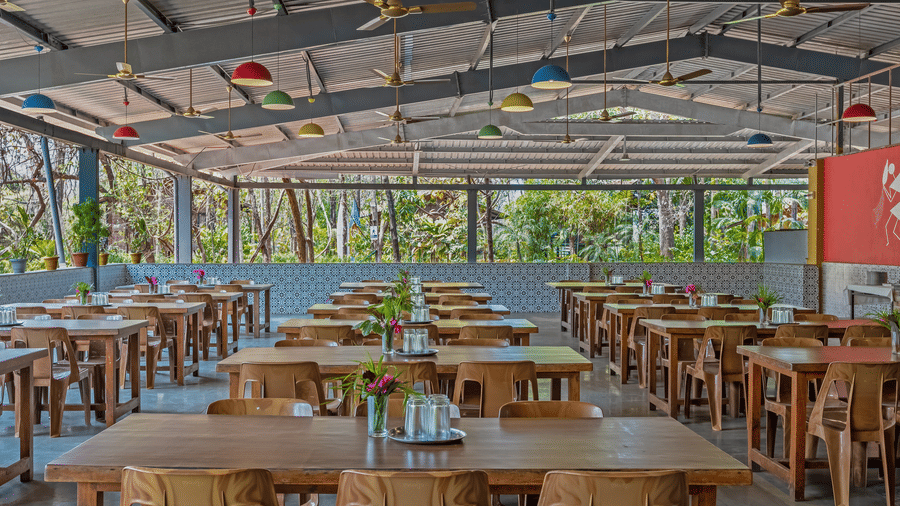 Wide-angle view of open-air resort dining hall with wooden furniture, transparent roof panels, colourful wall accents and lush greenery visible outside.