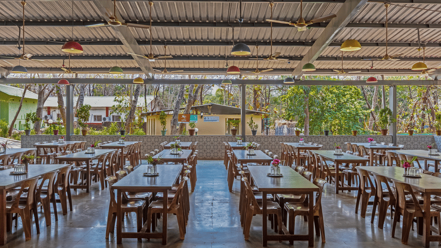 Open-air resort dining space with long wooden tables, transparent roof panels flooding natural light, green garden views and minimal decor.