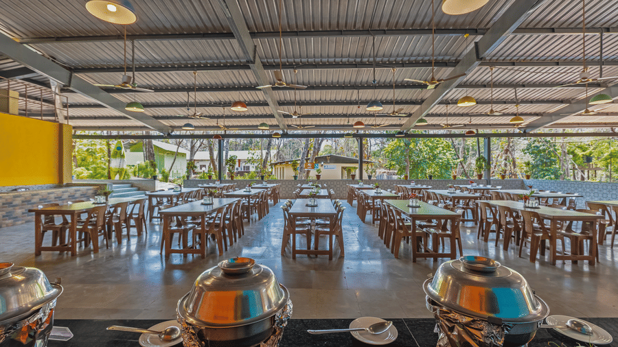 Spacious resort dining hall with wooden tables and benches, colorful multicolored walls, decorative pendant lights and open sides with garden views.