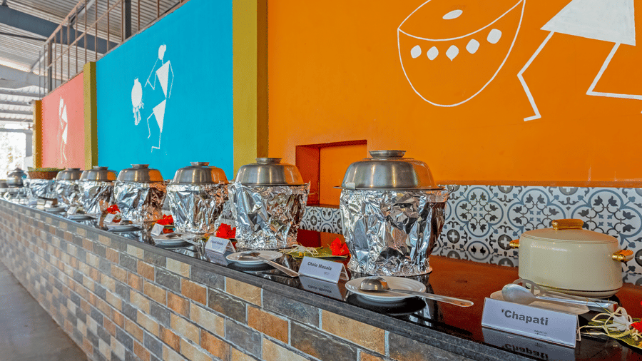 Close-up of resort buffet counter with food warmers, serving vessels, decorative tiles, and vibrant orange wall with tribal art illustrations in background.