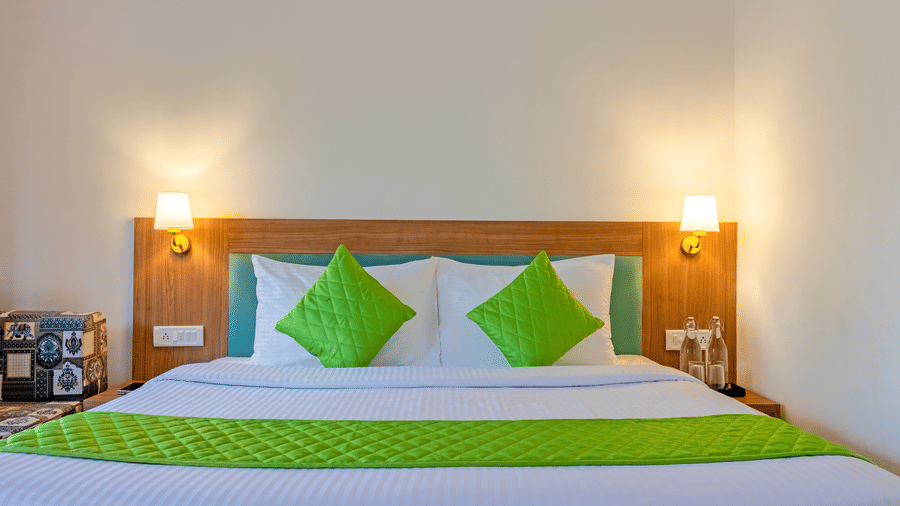 Close view of a double bed with green runner and cushions, wooden headboard, and warm bedside lighting.