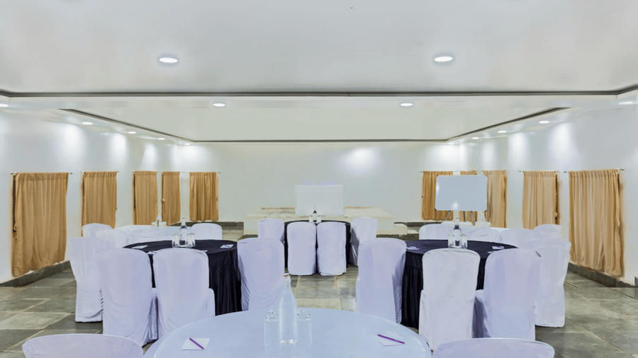 Elegant banquet hall with round tables draped in white and black covers, white chair covers, recessed ceiling lights and wooden panel wall backdrop.