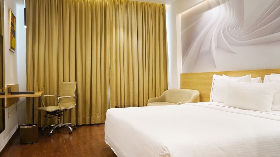 A corner view of a spacious guest room at Nemesia featuring a comfortable bed, a work desk, a seating area, and floor-to-ceiling curtains, creating a relaxing and inviting atmosphere.
