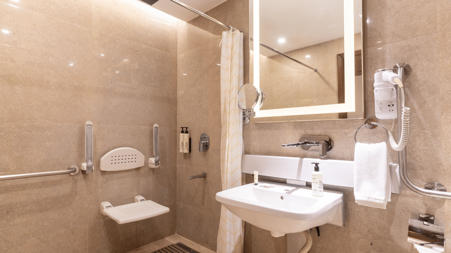 An accessible bathroom at Nirvana Sarovar Portico, Gorakhpur featuring a fold down shower seat, grab rails, a washbasin, and a mirror.  